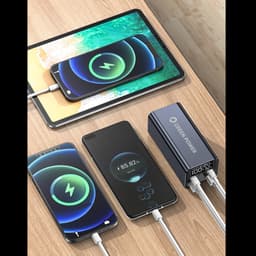 A black power bank with two cables, charging a tablet and two smartphones on a wooden surface.