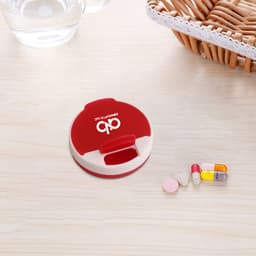 A round, red and cream pill box with a hinged lid, alongside assorted pills on a wooden surface.