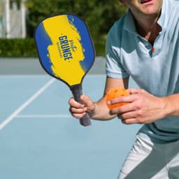 A wooden pickleball paddle in blue and yellow with an orange pickleball, held by a person on a court.