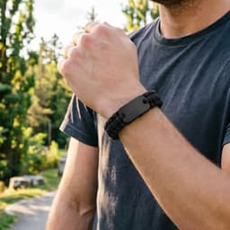 A black paracord bracelet featuring a metal plate, worn on a wrist amidst a natural outdoor setting.