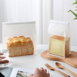 A clear sealing storage bag holds a loaf of bread beside another bag with a square loaf, both on a wooden surface.