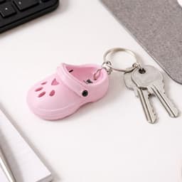 A pink paw shape clog shoe keyring attached to keys, placed on a desk near a notebook and computer.