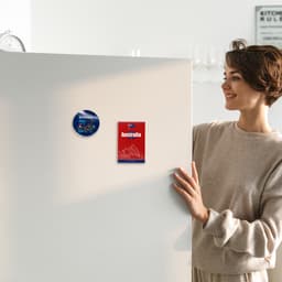 A woman in a light sweater stands next to a white fridge displaying two NFC acrylic magnets, one blue and one red.