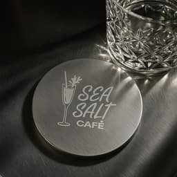 A round metal coaster with a shiny silver finish and a logo design. A glass sits beside it on a dark surface.