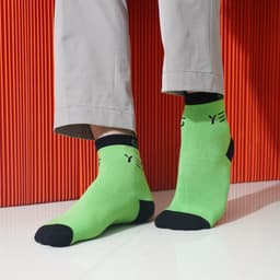 Bright green ankle socks with black accents and a logo, positioned on feet against a vibrant orange backdrop.