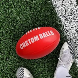 A red size 5 football with a white logo, resting on green artificial grass beside a shoe.