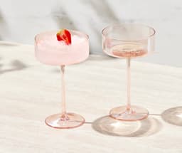 A set of two pink coupe glasses, one filled with a drink and garnished with a strawberry, displayed on a light surface.