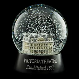 A snow globe featuring a detailed model of the Victoria Theatre, inside a transparent globe filled with sparkling snow.