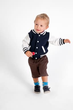 A child wearing a navy and white varsity jacket, brown shorts, and socks with blue accents, holding a small red object.
