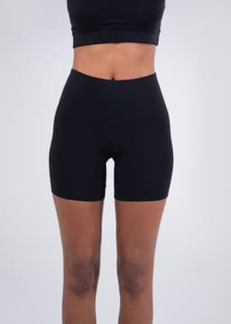 High-rise black legging shorts on a model, paired with grey sneakers. Features a brand logo on the waistband.