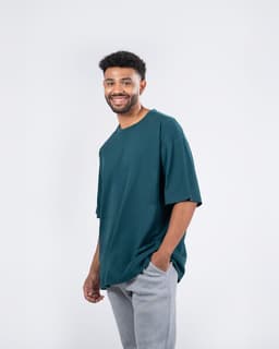 A man wearing a teal unisex oversize easy-fit tee and grey pants, smiling against a plain backdrop.