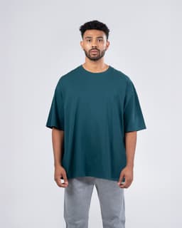 A man wearing a dark teal unisex oversize easy-fit tee, paired with grey trousers, standing against a plain background.