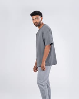 A grey unisex oversize easyfit tee displayed on a model, paired with light grey trousers, featuring a brand logo.