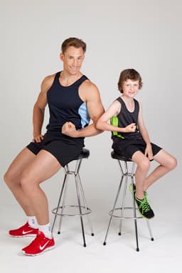 A man and a boy pose on stools, wearing black and blue singlets, showcasing a sporty style. The man has red shoes and the boy wears green shoes.