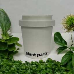 A bamboo coffee cup with a hard lid in a light grey colour, featuring a logo and surrounded by greenery.