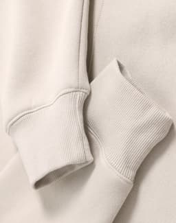 A close-up of a beige oversized hoodie featuring ribbed cuffs and a soft fleece texture. It has a logo.