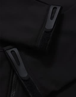 Close-up of a black softshell jacket featuring zippers and velcro details. The jacket has a logo on it.