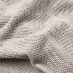 A textured blanket in soft beige tones, featuring a woven pattern and slight sheen, with a visible logo.