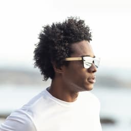 Sunglasses with reflective lenses and wooden frames, worn by a person with curly hair, against a scenic outdoor background.