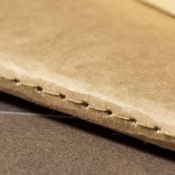 A close-up of a beige notebook's spine with visible stitching on a brown background. The notebook has a subtle texture.