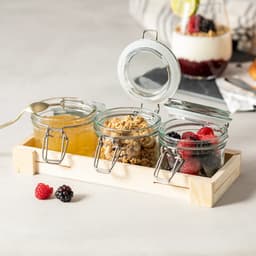 Three glass jars with metal clips, containing honey, granola, and mixed berries, placed on a wooden tray.