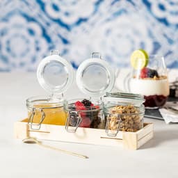 A set of three glass jars with metal clips, containing fruit, berries, and granola, presented on a wooden tray.