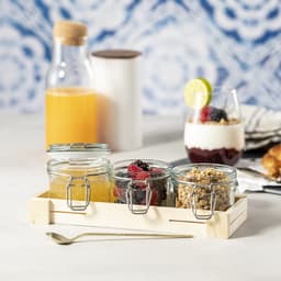 A wooden tray holds three glass jars filled with granola and mixed berries, alongside a drink bottle and a dessert glass.