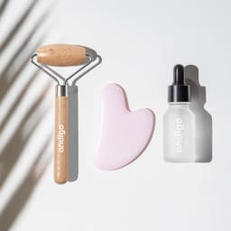 A pink face massager, a wooden roller, and a dropper bottle on a light surface. The items feature a logo.