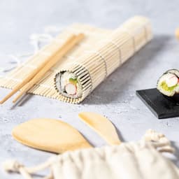 A sushi rolling mat, sushi rolls on a black plate, and wooden utensils. The mat features a bamboo design.