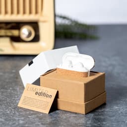 A pair of white earbuds in a brown and white box, set against a textured grey surface. The packaging features a logo.
