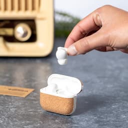 A pair of white earphones being held above a cork case, set against a grey surface and a vintage appliance in the background.