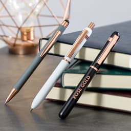 A collection of three metal pens in grey, white, and rose gold, resting on stacked books with a geometric lamp in the background.
