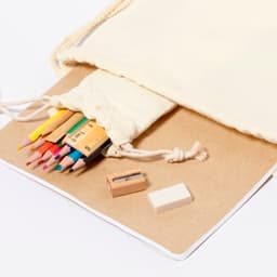 A cream drawstring pouch containing coloured pencils, a sharpener, and an eraser on a craft paper surface.