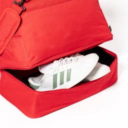 A red duffle bag with a shoe compartment containing white sneakers with green stripes and a branding logo.