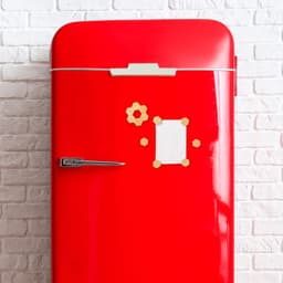 A red fridge with a white paper and decorative magnets attached to its surface against a brick wall.