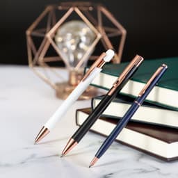 Three metal pens in white, black, and navy, featuring rose gold accents, positioned beside stacked books.