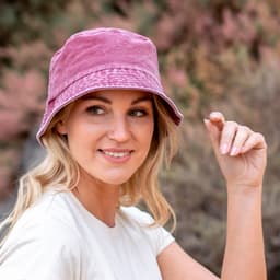A woman wearing a pink bucket hat, smiling while standing against a blurred natural background. The hat features a logo.
