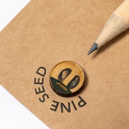 A grey-lead pencil with a sharpened tip resting on a brown card featuring a circular seed design and a logo.