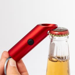 A red keyring bottle opener with a rounded top, featuring a button, attached to a glass bottle.