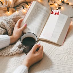 A warm drink in a black mug beside an open book with a decorative bookmark, all set on a cozy blanket.