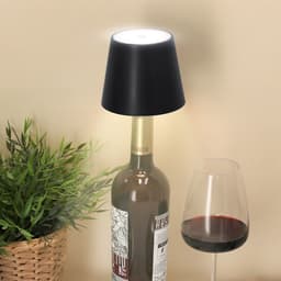 A black lamp sits atop a wine bottle, illuminated, beside a glass of red wine and a potted plant.