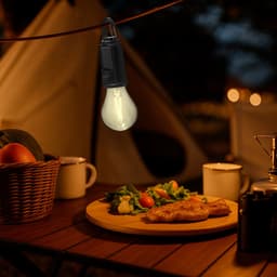 A warm white hanging lamp illuminates a campsite table with a plate of food, surrounded by a basket and drinks.