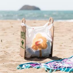 A cardboard bag holder in sand with a white trash bag featuring a flower design, by the beach near water.