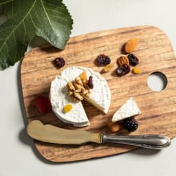 A wooden cutting board featuring a round cheese wheel, nuts, dried fruits, and a silver knife, with a leaf in the background.