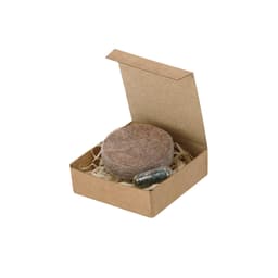 A round compressed seed disk in a small cardboard box, accompanied by a capsule, sitting on shredded paper.