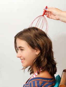 A red and silver head massager is being used on a girl’s head, providing relaxation. The background is neutral.