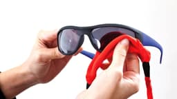 A pair of sunglasses held by hands, featuring a red and black multipurpose glasses strap.