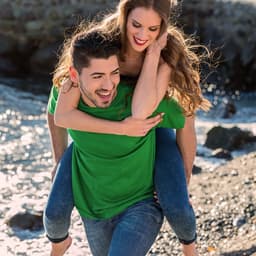 A green premium adult t-shirt displayed on a couple on a beach, with one person giving a piggyback ride.