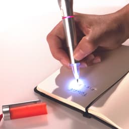 A silver and red pen with a built-in torch lights up while writing in a notebook, alongside an orange cap.