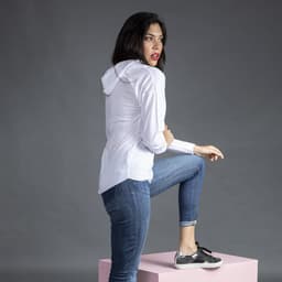 A woman in a white Yuk T-Shirt with a hood, wearing blue jeans and black shoes, posed on a pink pedestal.